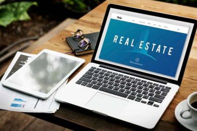 Laptop displaying 'REAL ESTATE' on screen, with tablet and coffee.