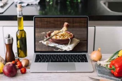 Laptop displaying pasta recipe with fresh ingredients on counter.